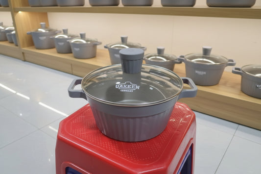 Stainless steel pot with a glass lid on a red stand, branded 'Uakeen Germany', in a cluttered indoor setting.