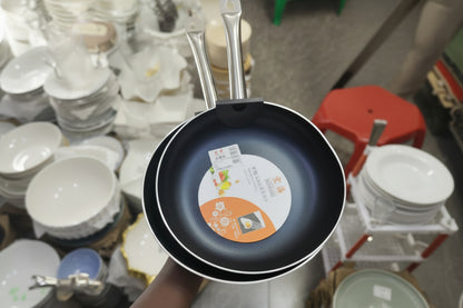 Black frying pan with a brand label held by a person on a cluttered floor.