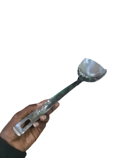 Person holding a large spoon in front of a cluttered shelf with various items.