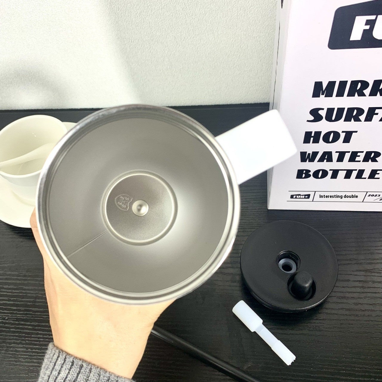 Metallic hot water bottle with black cap held by a hand, with a branded box in the background.