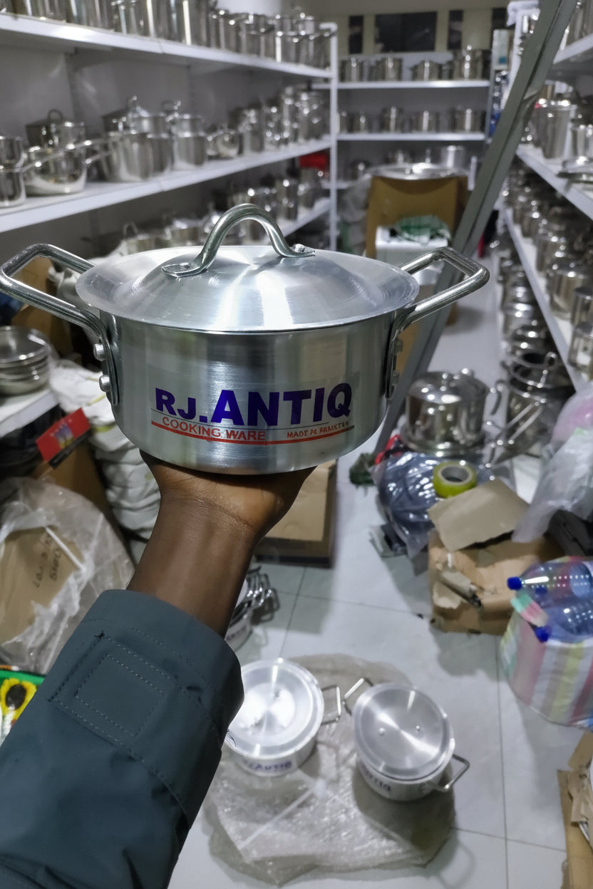 Stainless steel pot with 'RJ Antigua' branding held by a person in a cluttered indoor setting.