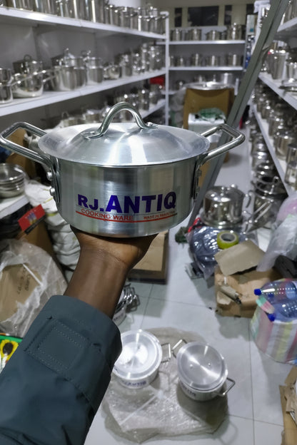 Stainless steel pot with 'RJ Antigua' branding held by a person in a cluttered indoor setting.