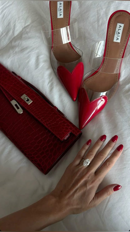 Red high-heeled shoes, red handbag, and a hand with red nails and a ring on a white surface.