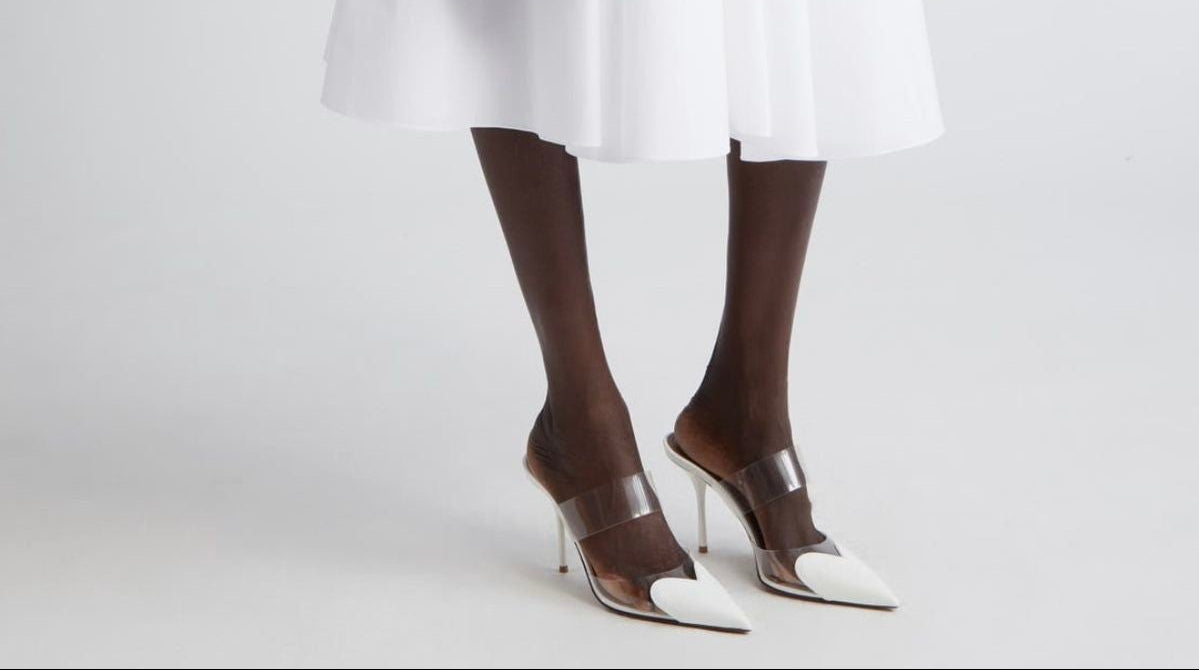 Person wearing a white dress with a black belt and holding a heart-shaped bag on a light gray background