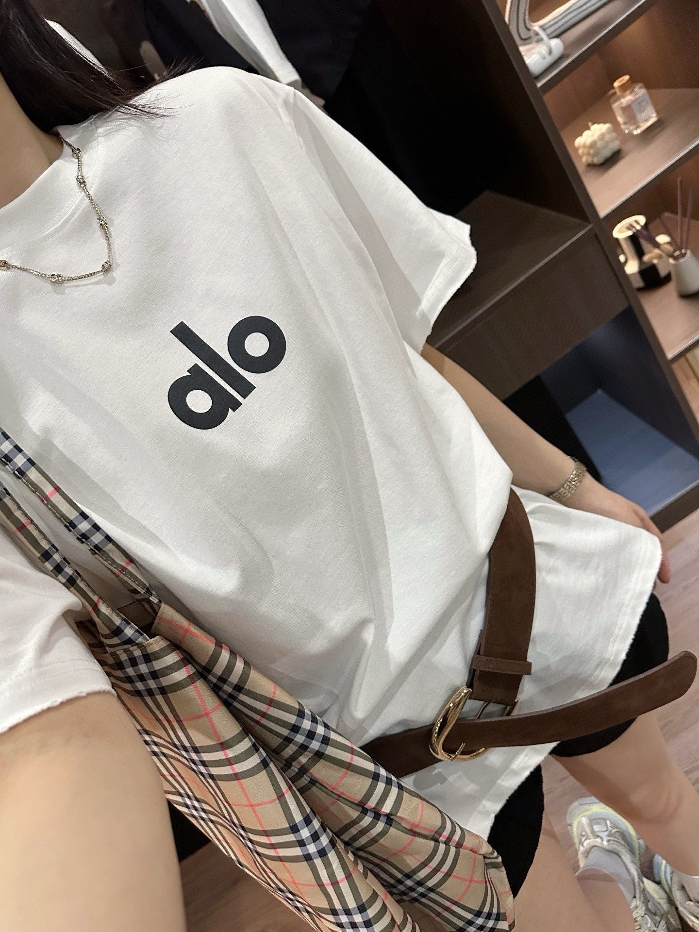 Person wearing a white shirt with 'alo' logo and plaid scarf, sitting indoors.