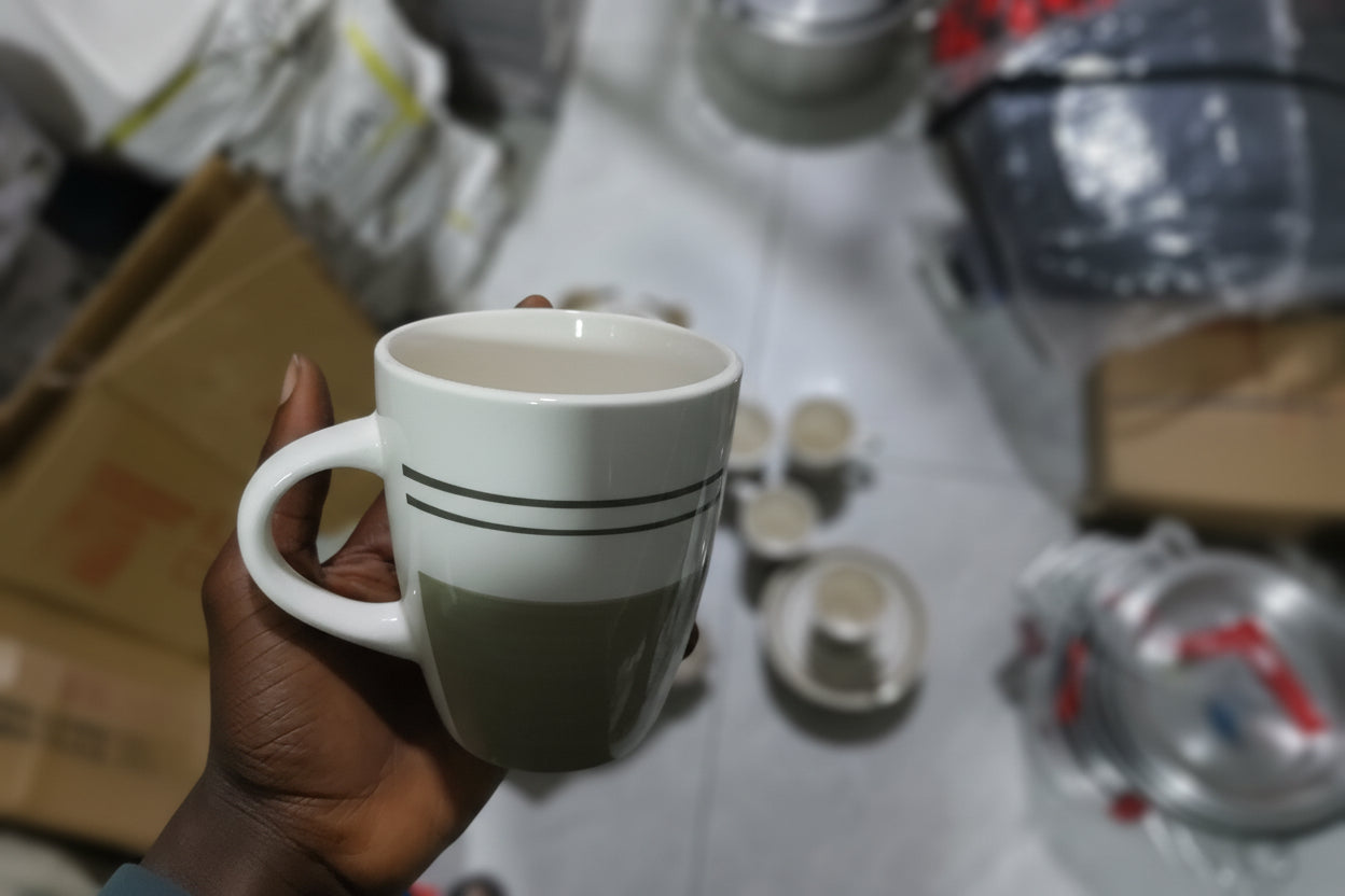 Hand holding a white mug with a gray lid in a cluttered room.