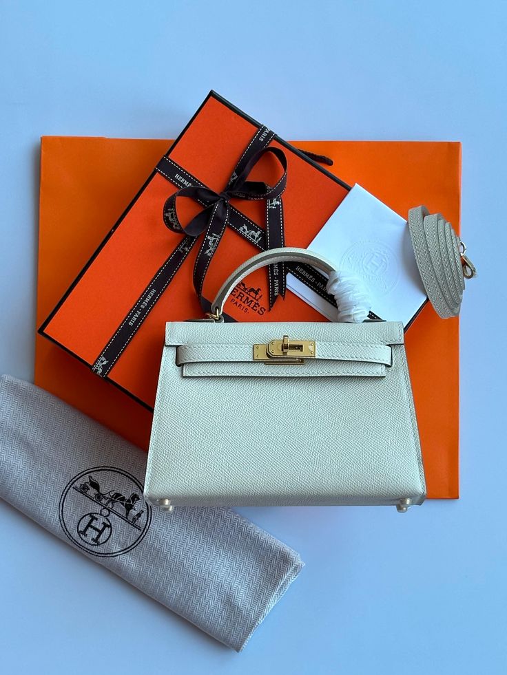 Light gray handbag with gold hardware on an orange Hermes box, with a gray cloth and black logo card.
