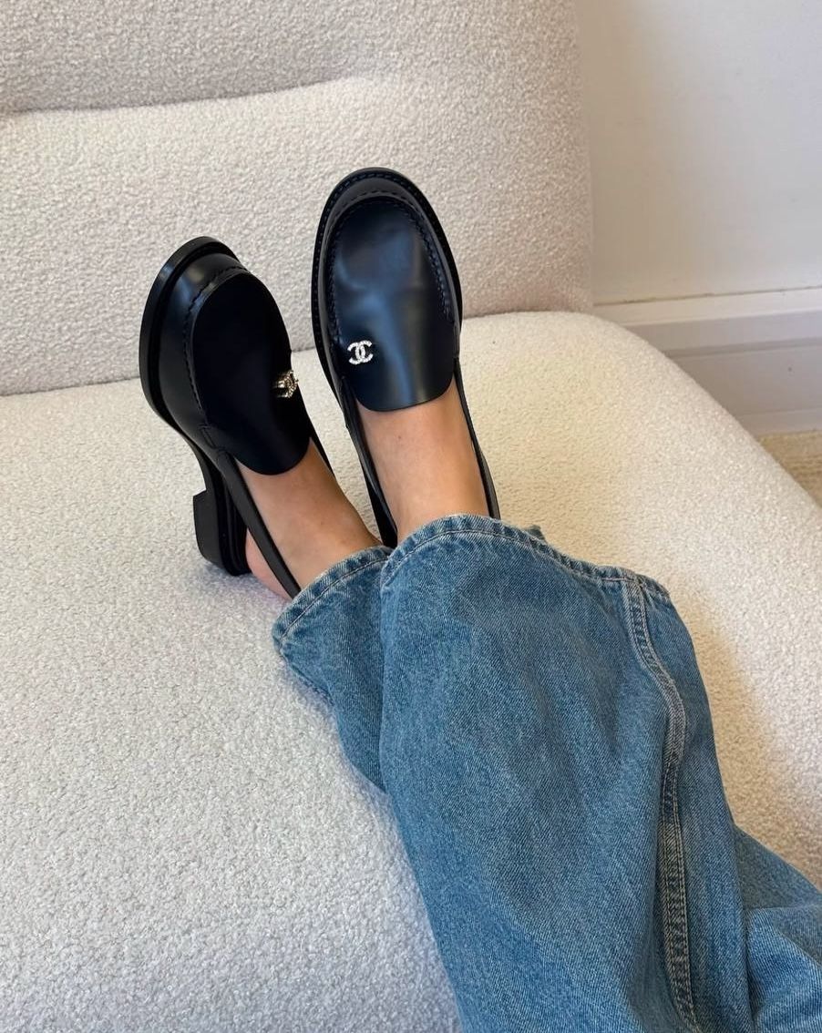 Black shoes with a visible brand logo worn by a person sitting on a light-colored couch.