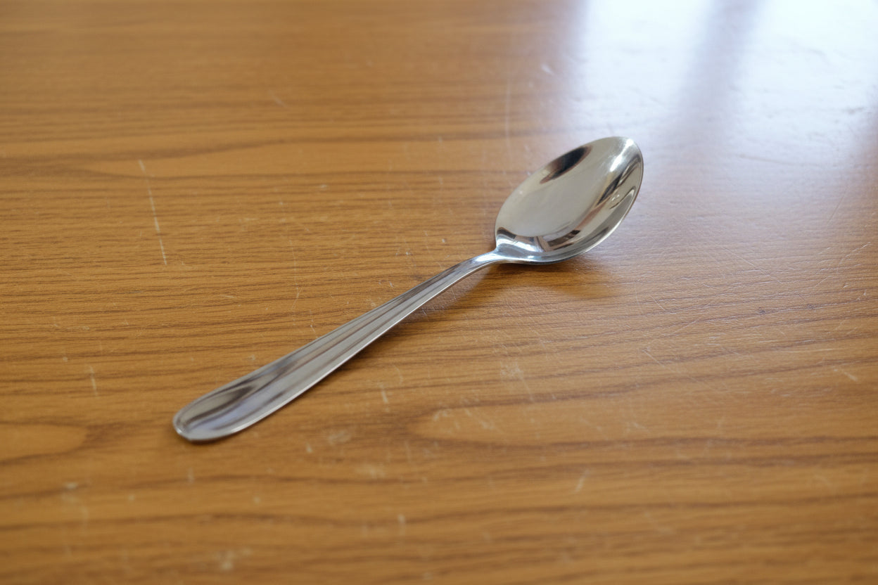 Hand holding a silver spoon with a blurred background of stacked white containers and cardboard boxes.