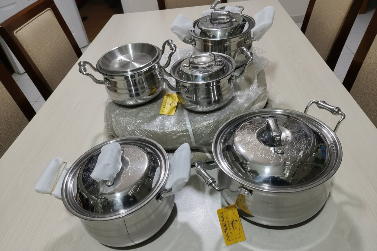 Set of stainless steel pots and pans on a tiled floor with colorful packaging in the background.