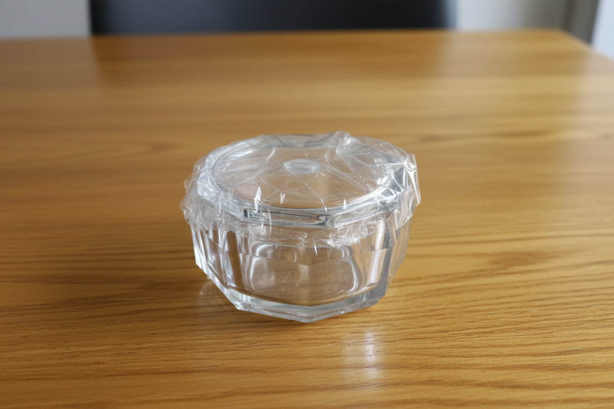 Hand holding a clear plastic container with a lid, surrounded by stacked items on a tiled floor.
