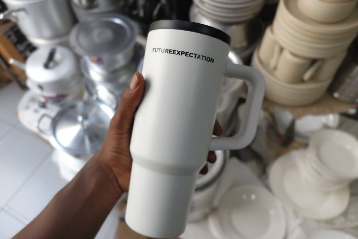 White insulated mug with black handle and 'FUTUREEXPECTATION' branding held by a hand against a blurred background.