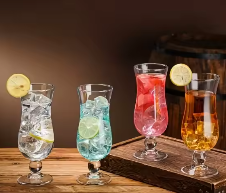 Four glasses of different colored drinks with ice and lemon slices on a wooden surface.
